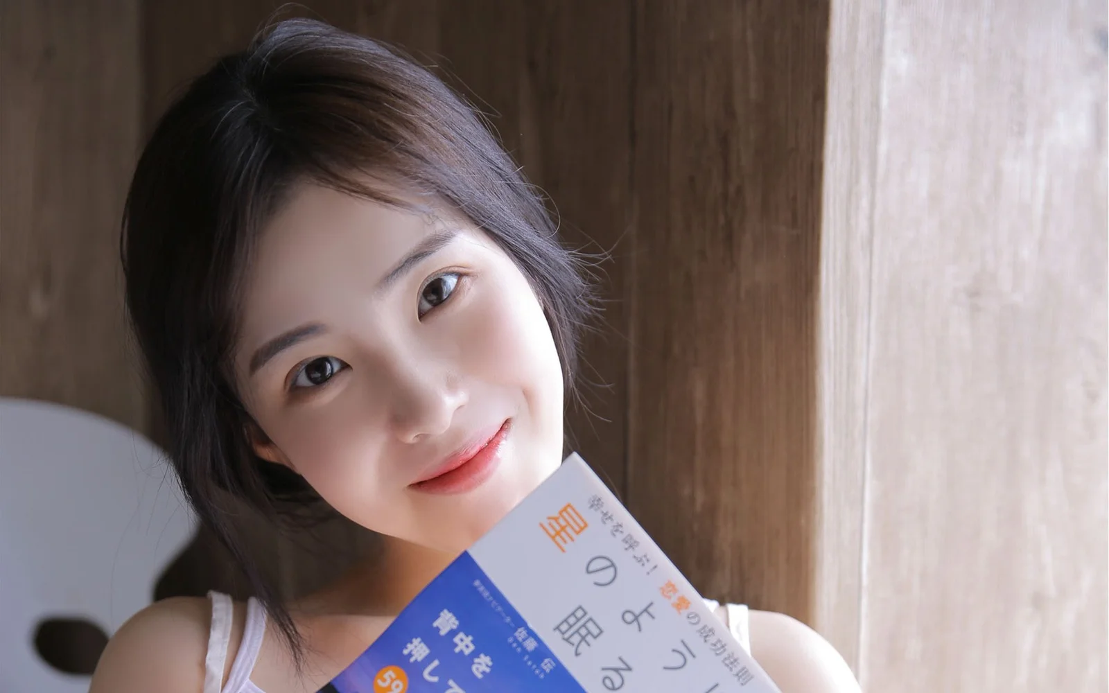 成人女性が木目調の室内を背景に本を持ってカメラに微笑みかけている。