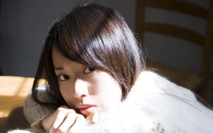 茶色の髪の女性が白いニットを着て、日差しの中でカメラを見つめています。