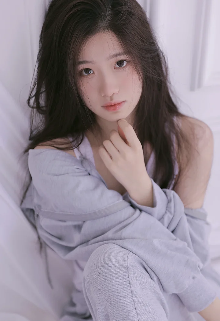 一名长发女性在柔和的淡紫色室内背景前抱膝坐着，手指轻触下巴并看向镜头。