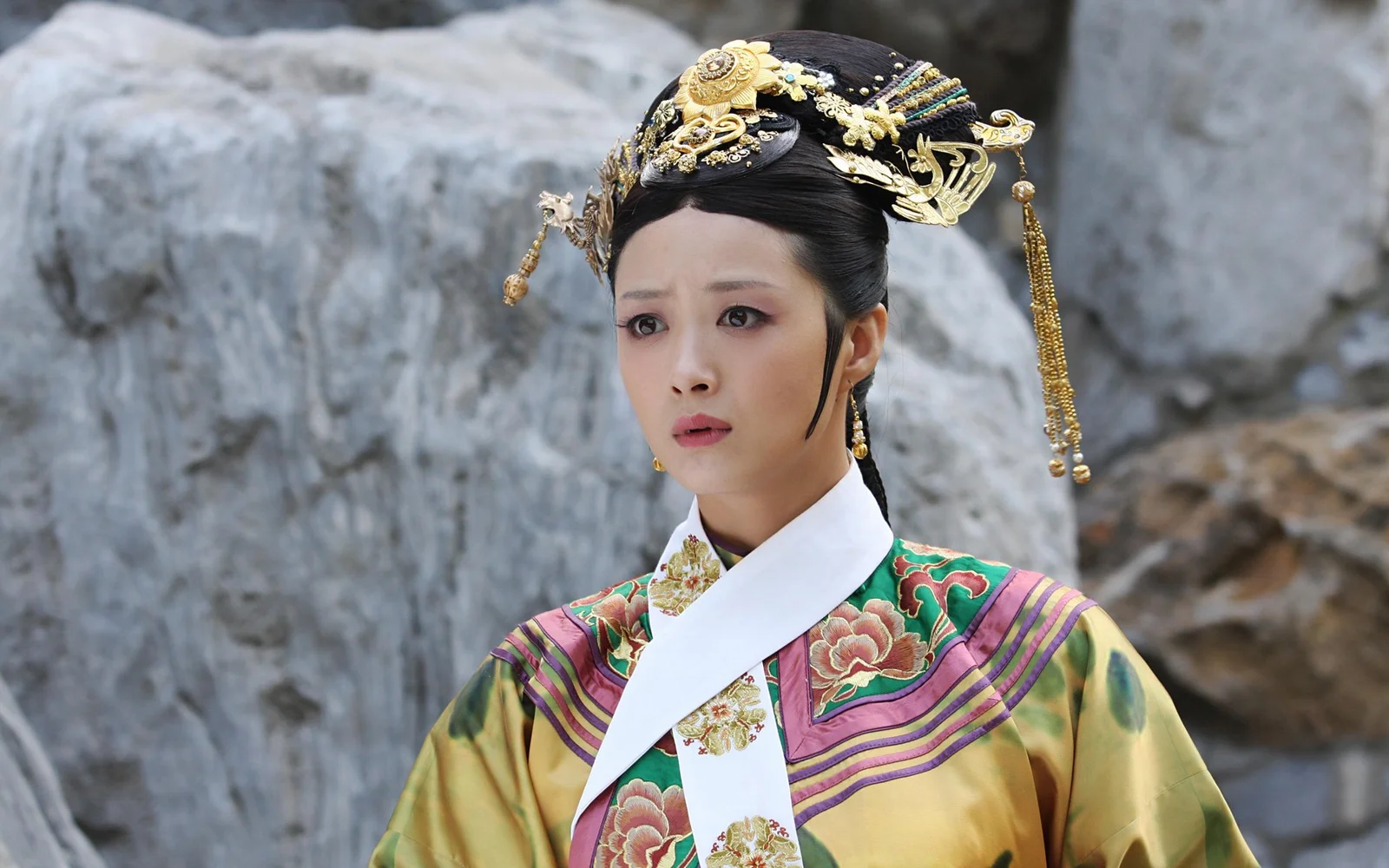 화려한 전통 의상과 장식용 머리 장식을 한 성인 여성이 바위 배경 앞에 서 있습니다.