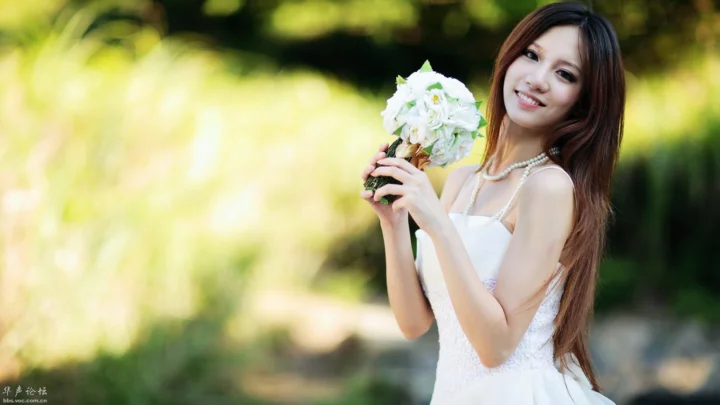 一位成年女性在绿意盎然的户外手捧白色花束微笑面对镜头。