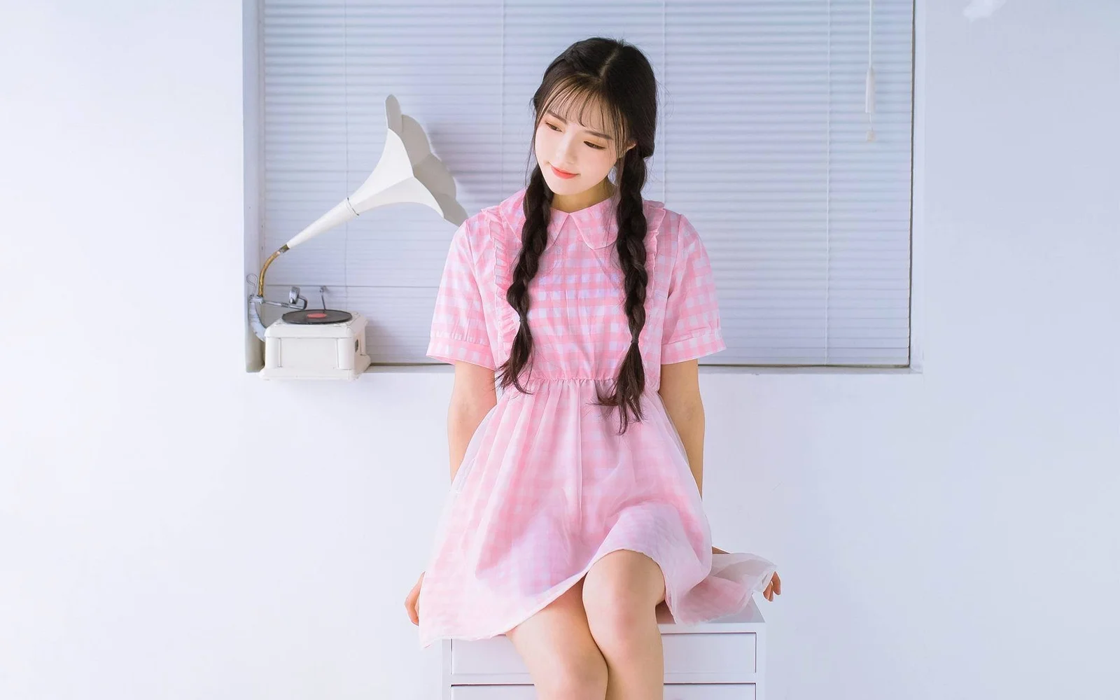 ピンクのワンピースを着た三つ編みの女性が、室内の窓辺に座っています。