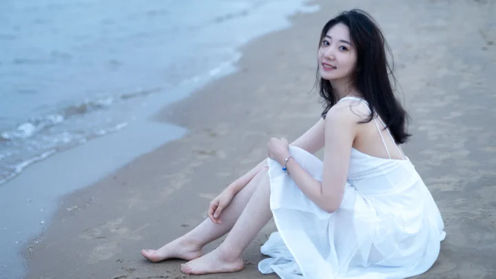 흰색 원피스를 입은 여성이 잔잔한 바다와 모래사장을 배경으로 해변에 앉아 있습니다.