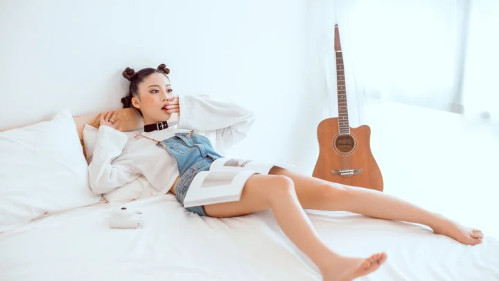一名成年女性穿着白色上衣和牛仔短裤躺在白色床上，身旁放着一本书，背景靠着一把木吉他。
