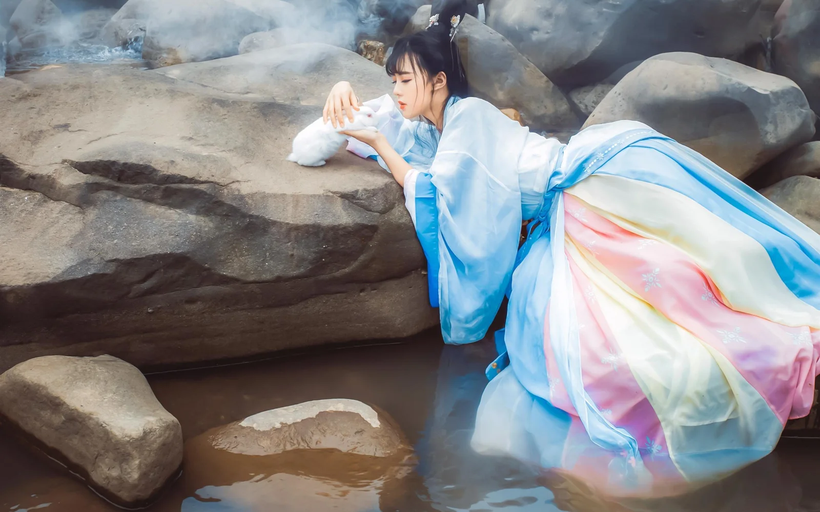연한 파란색 전통 의상을 입은 여성이 얕은 물가의 바위 사이에서 쉬고 있다.