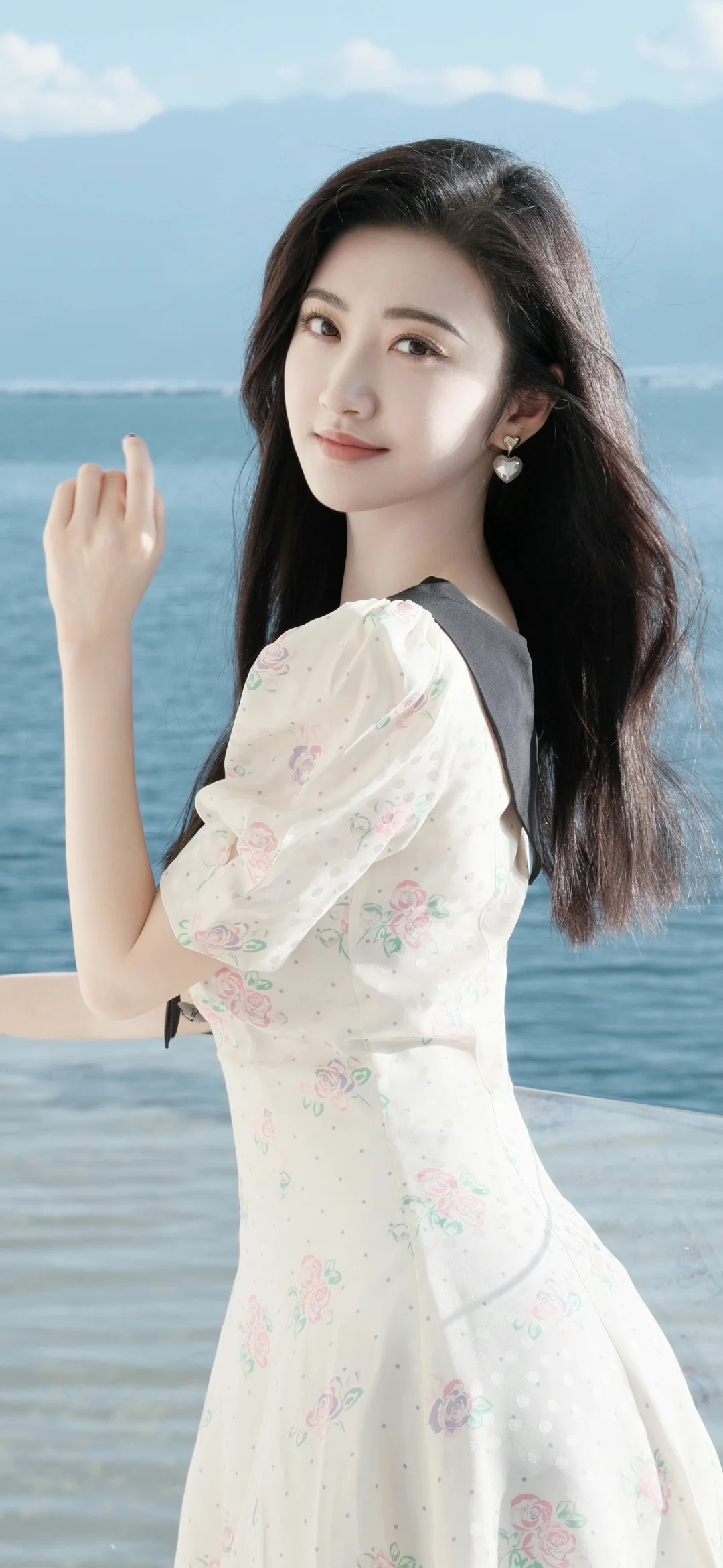 긴 머리의 여성이 흰색 무늬 원피스를 입고 바닷가에 서 있습니다.