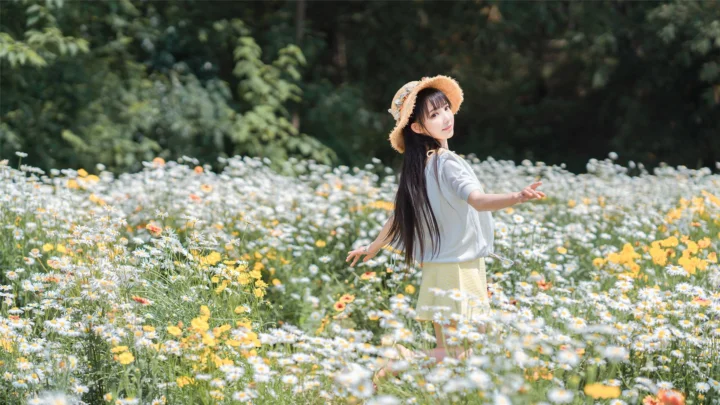 한 여성이 활짝 핀 야생화 들판에 서 있고 뒤로 울창한 푸른 숲이 펼쳐져 있습니다.