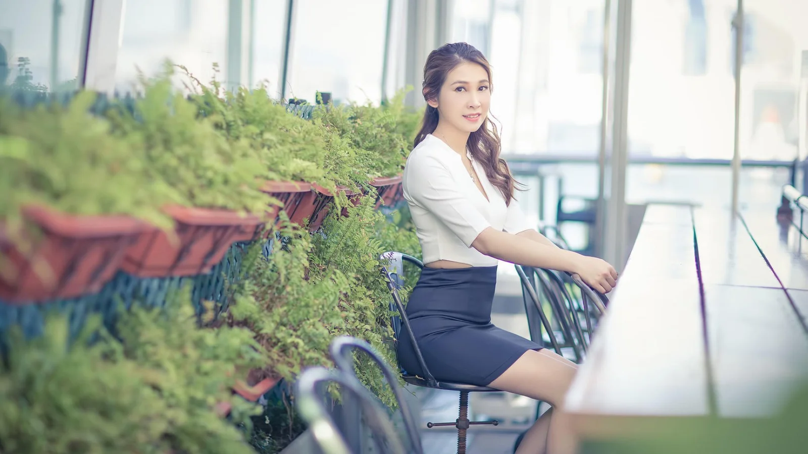 一名成年女性坐在明亮的办公室窗边，身旁摆放着多盆绿色植物。
