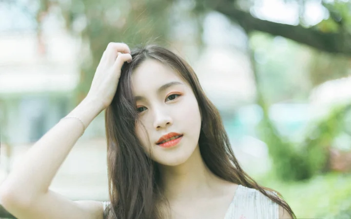 一名长发女性在户外绿意背景前轻抚头发并看向镜头。