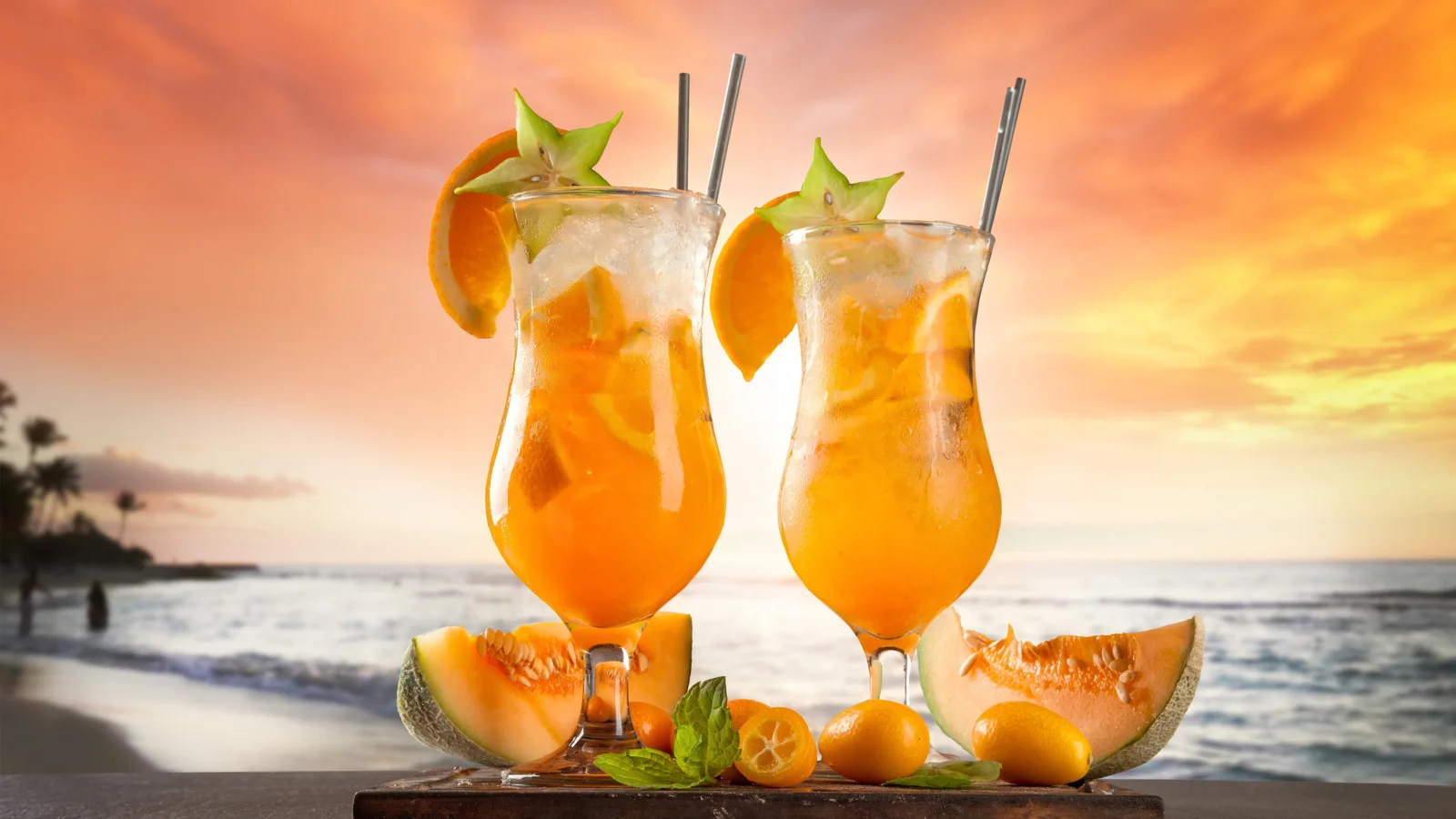 2杯のオレンジ色のドリンクとフルーツを手前に、海辺と鮮やかな夕焼け空が広がっています。