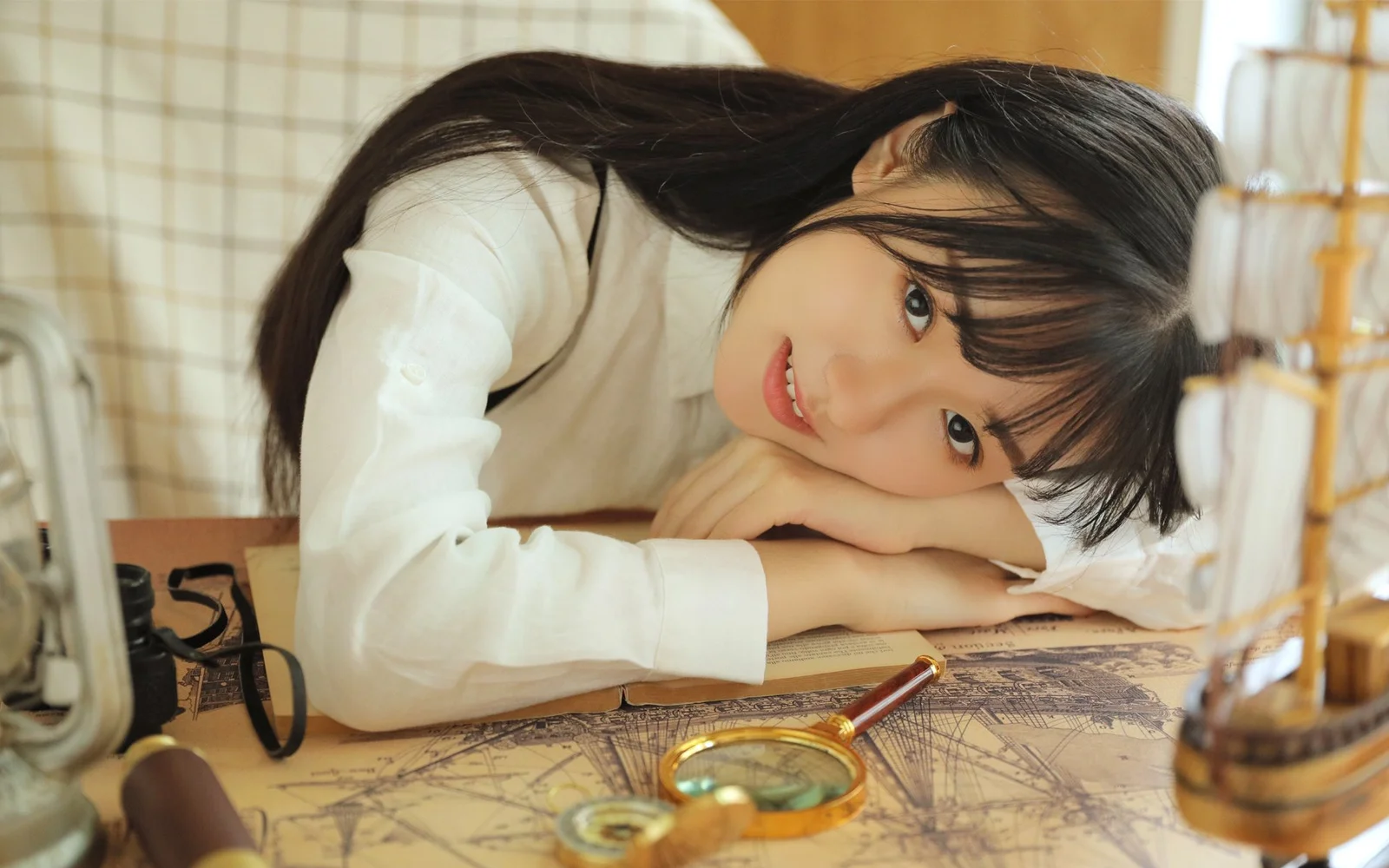 白いトップスを着た長い髪の女性が、地図と虫眼鏡の置かれたテーブルに身を預けています。