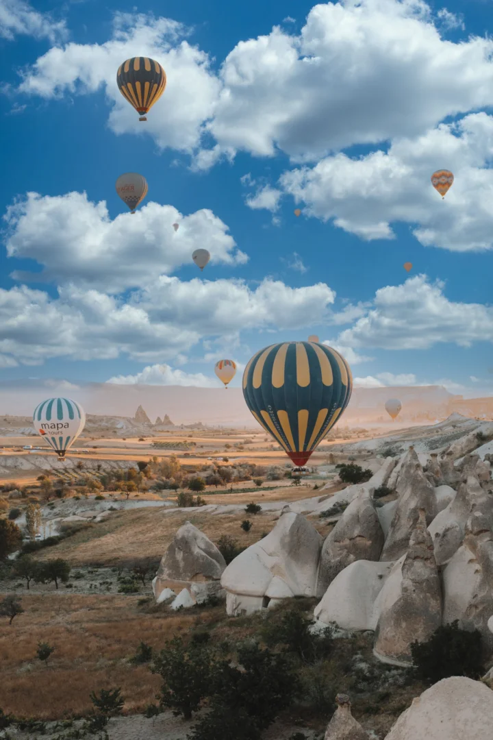 蓝天白云下，多只热气球飘浮在岩石地貌与干燥山谷上空。