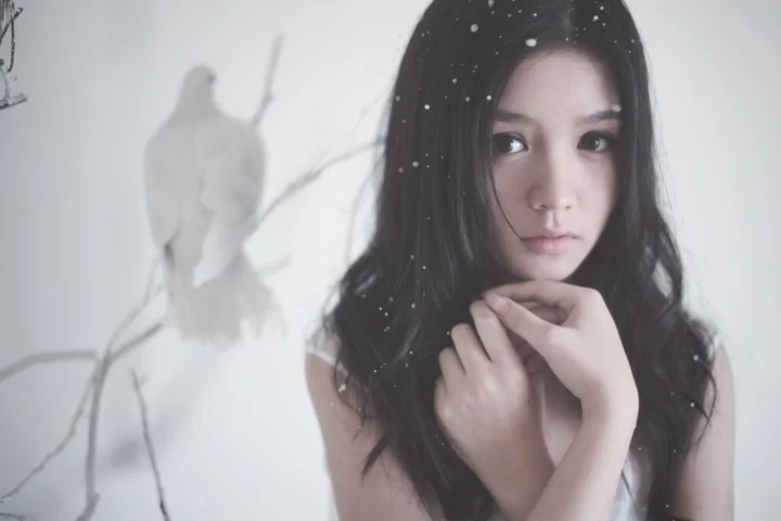 一名黑发女子在白色背景前凝视镜头，发丝和画面中点缀着细小白色雪点。