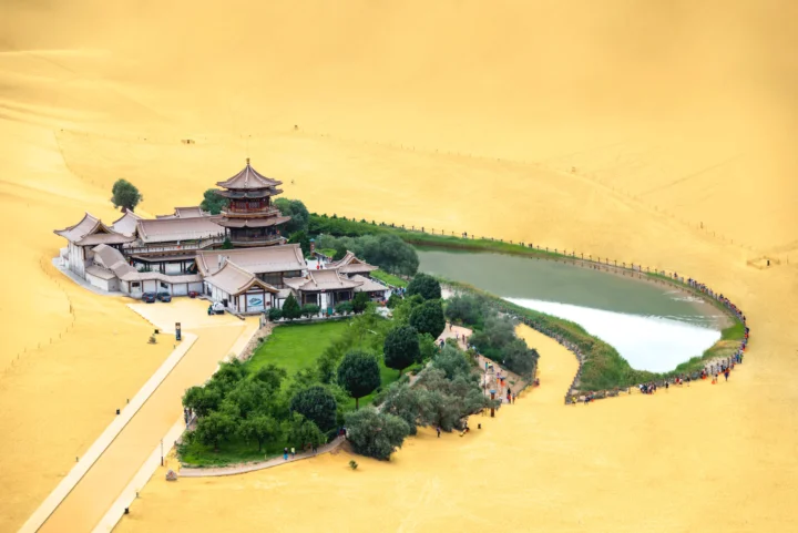 金色沙漠中的月牙形湖泊旁坐落着一组古建筑和绿洲植被。