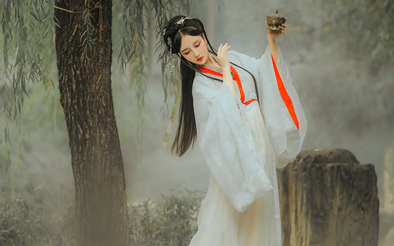 一名身穿白色传统服饰的女子站在柳树和树桩之间的户外朦胧景色中。