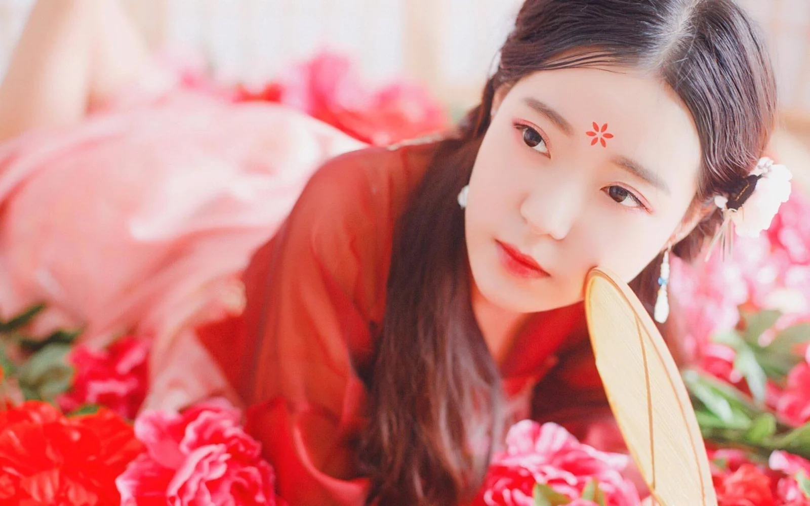 赤い服の女性が花々の中で扇を持ち、カメラに顔を向けています。
