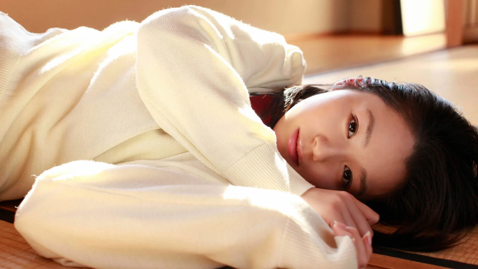 밝은 색 스웨터를 입은 검은 머리 여성이 나무 바닥에 누워 카메라를 바라보며 부드럽게 미소 짓고 있습니다.