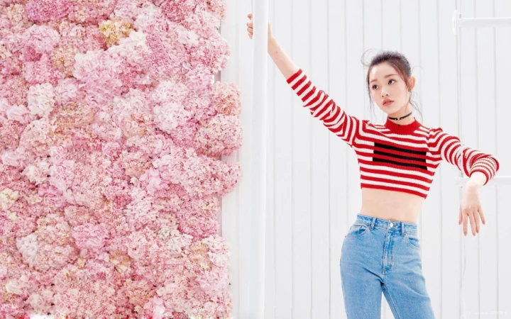 一名穿红白条纹短款毛衣和牛仔裤的成年女性站在粉色花墙与白色墙板前。