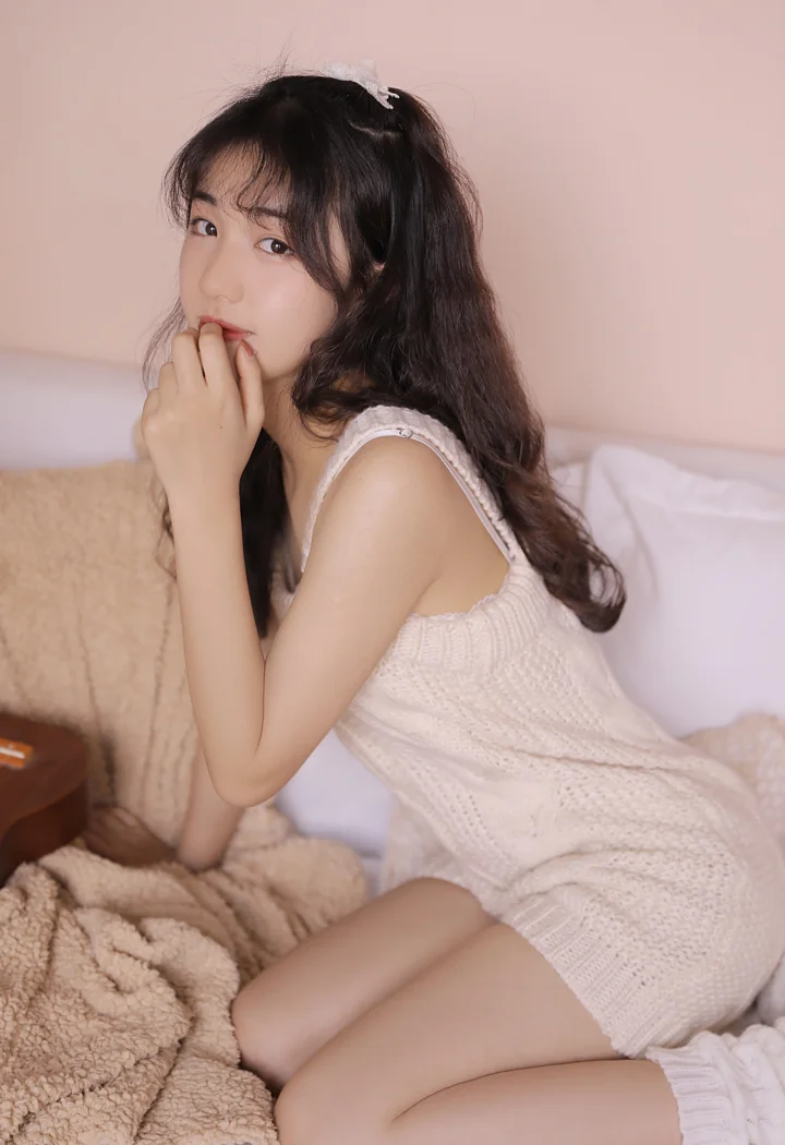 밝은 색상의 니트 원피스를 입은 여성이 침대에 앉아 카메라를 바라보며 포즈를 취하고 있다.
