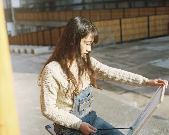 クリーム色のセーターとデニムのオーバーオールを着た成人女性が、日なたの屋外でイーゼルのそばに座っています。