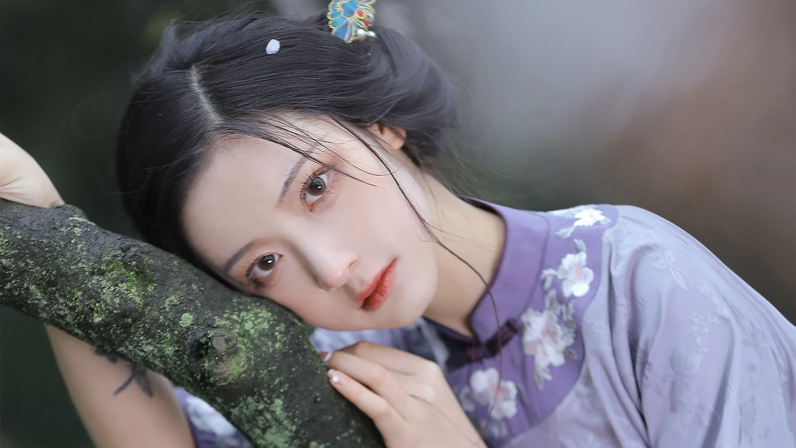 一名身穿淡紫色花紋服飾的女子將頭靠在覆有苔蘚的樹枝旁。