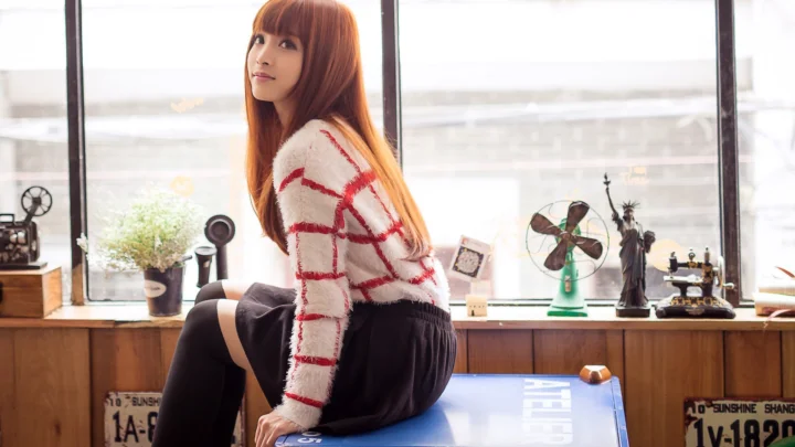 一名紅髮女性穿著白色紅紋毛衣和黑色長襪坐在室內窗邊。