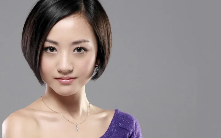 一名短发女性面对镜头，身穿紫色上衣，背景为纯灰色。