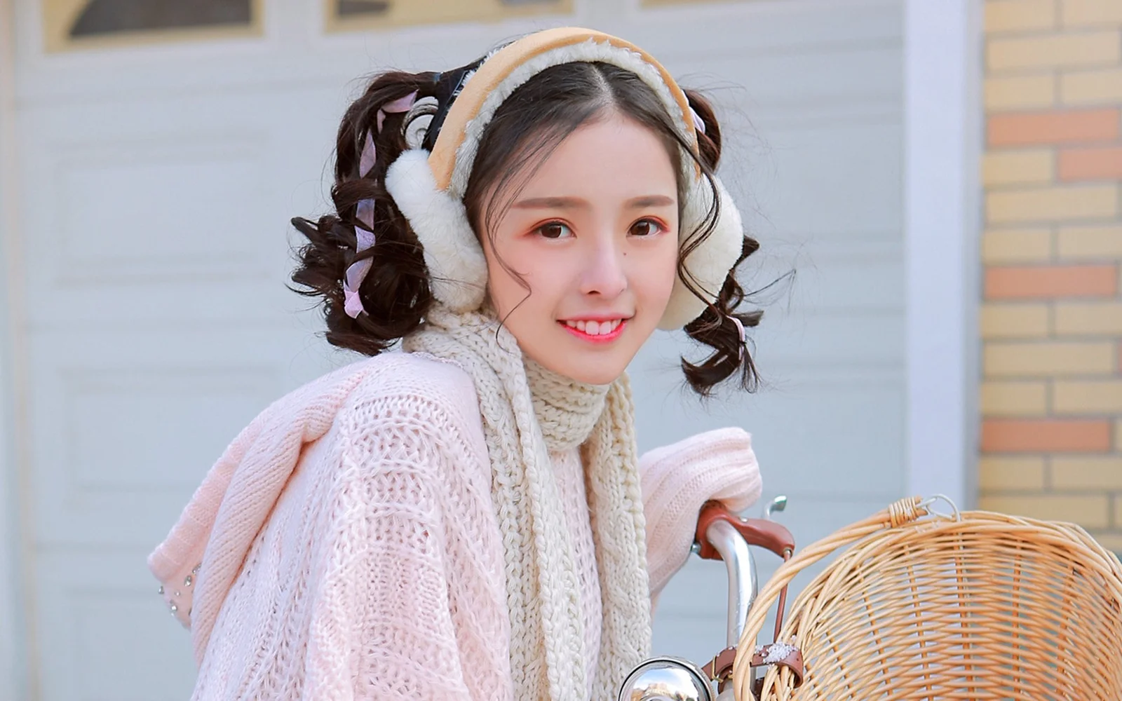 귀마개와 목도리를 착용한 젊은 여성이 밝은 색 니트 옷을 입고 건물 밖에 서 있습니다.