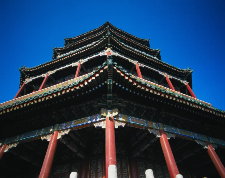 镜头仰拍一座饰有彩绘屋檐和红色立柱的中式古建筑，背景是晴朗的深蓝色天空。