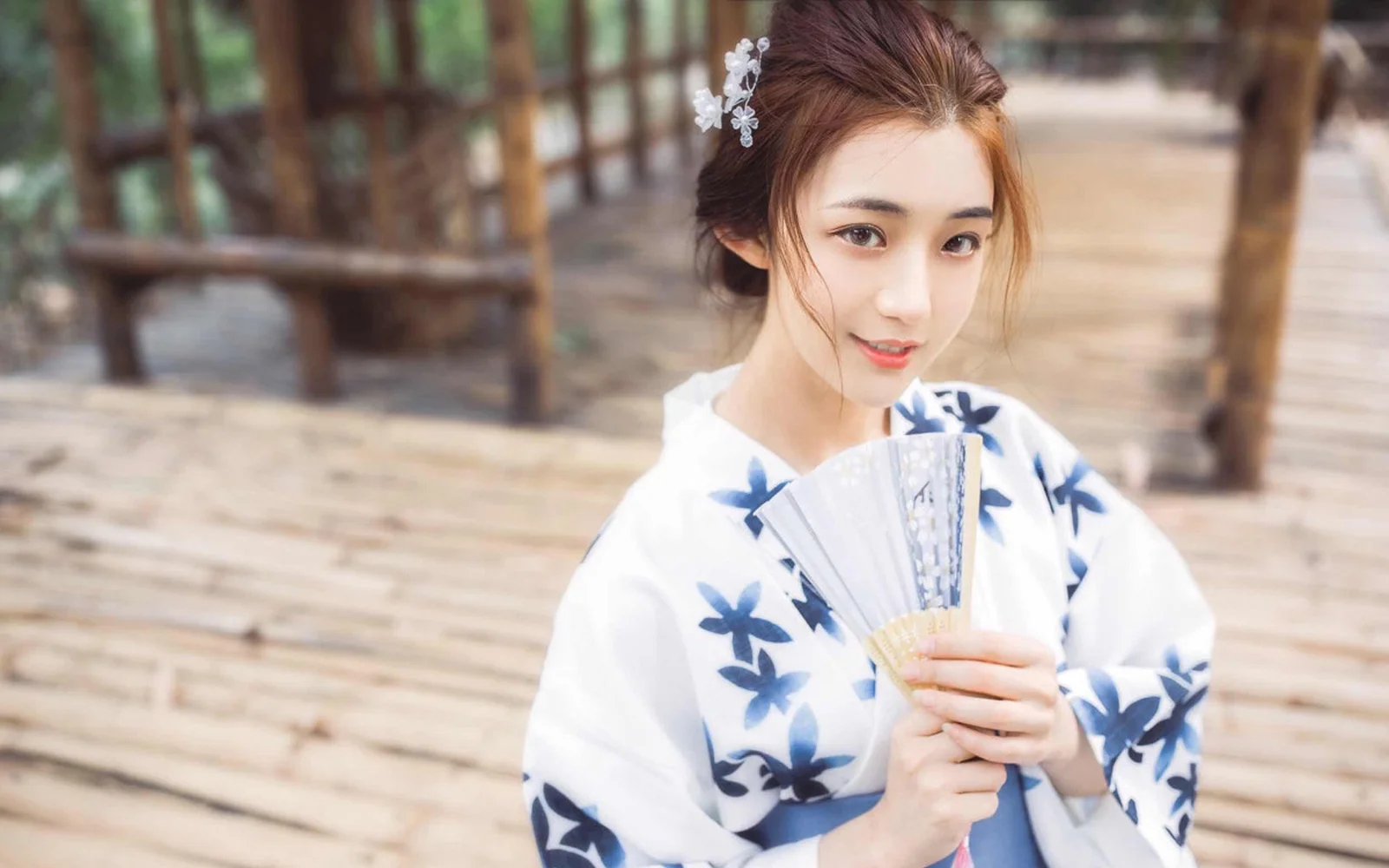 一名身穿白色藍花傳統服飾的女子在木質步道上手持摺扇。