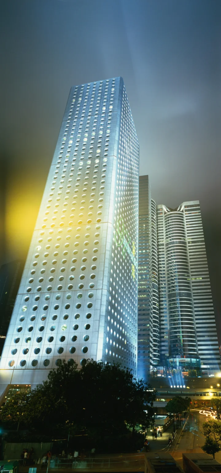 夜色中几座现代高层建筑被灯光照亮，前景可见树木和城市道路。