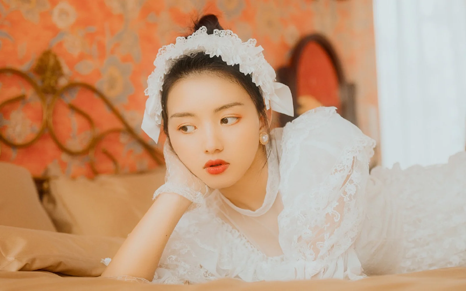 一名戴着白色蕾丝头饰、身穿白色蕾丝服装的女性倚靠在手边，背景是暖色图案墙面。