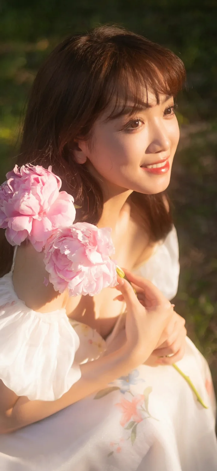 一名成年女子在户外阳光下手持粉色花朵，身穿白色连衣裙。
