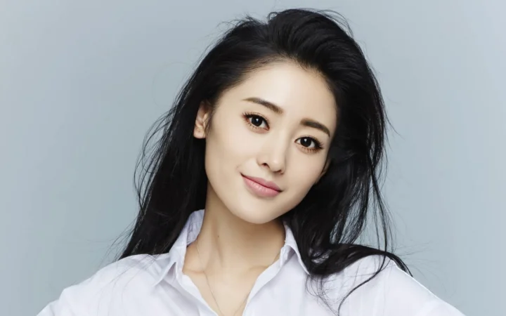 검은 머리의 여성이 흰 셔츠를 입고 연한 회색 배경 앞에서 카메라를 향해 미소 짓고 있다.