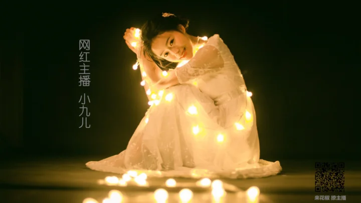 一名身穿白色连衣裙的女子坐在昏暗背景前，身上和地面散落着温暖的灯光。