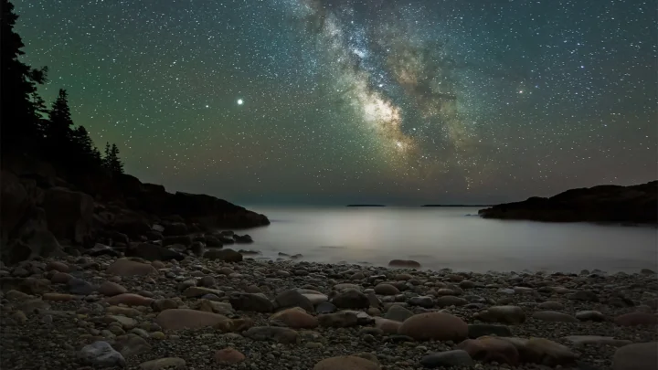 银河横跨繁星密布的夜空，下方是宁静海面与布满圆石的海岸。