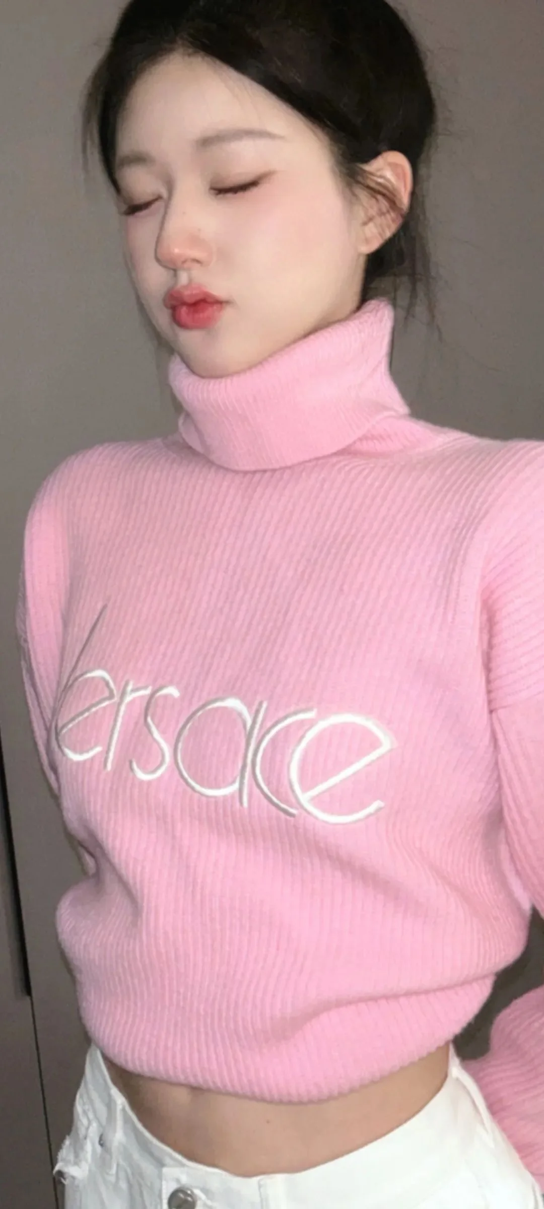 目を閉じた女性が室内でピンクのタートルネックセーターを着ています。