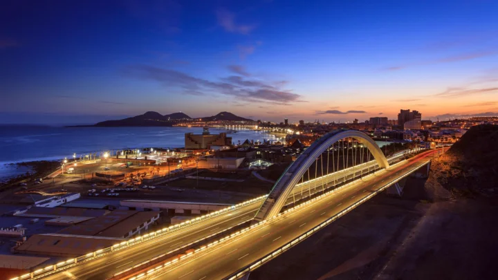 暮色中的海滨城市灯火初上，一座拱桥横跨道路，远处是海岸与城市建筑。