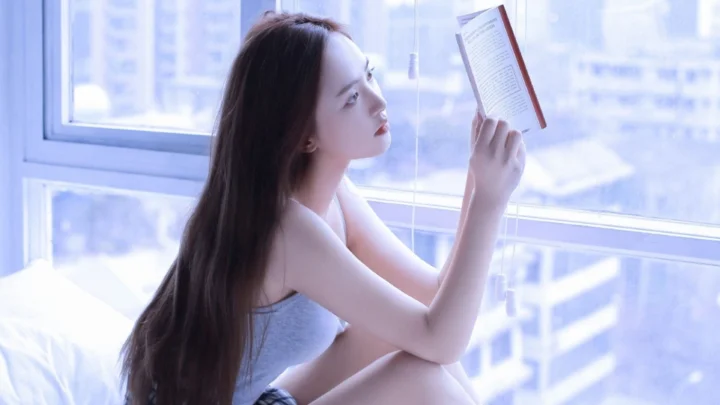 一名女性坐在窗边，手持书本阅读，明亮的窗光映照出室内的柔和氛围。