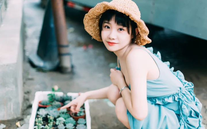一名戴草帽、穿藍色連身裙的女性在戶外花盆旁回頭看向鏡頭。