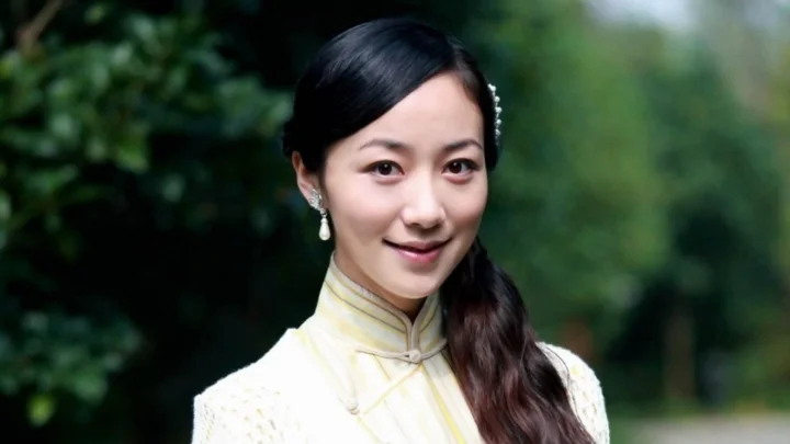 밝은 색상의 하이넥 의상을 입은 성인 여성이 푸른 야외 배경 앞에서 카메라를 향해 미소 짓고 있습니다.