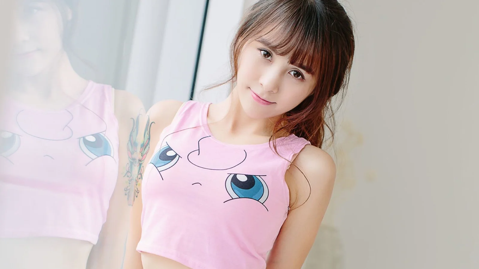 분홍색 만화 무늬 상의를 입은 미소 짓는 여성이 거울 옆에 서 있으며 화면에 그녀의 모습이 비칩니다.