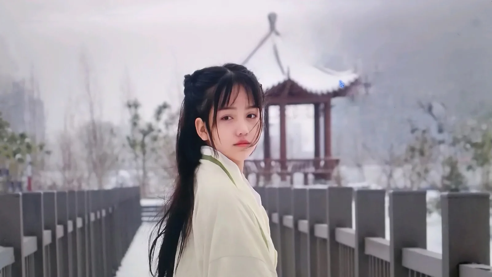 一名长发女子站在桥上回头望向镜头，背景可见朦胧的亭子和树木。