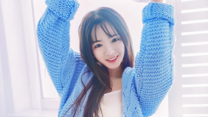 파란 니트 스웨터를 입은 긴 머리 여성이 밝은 창가에서 두 팔을 들고 미소 짓고 있다.
