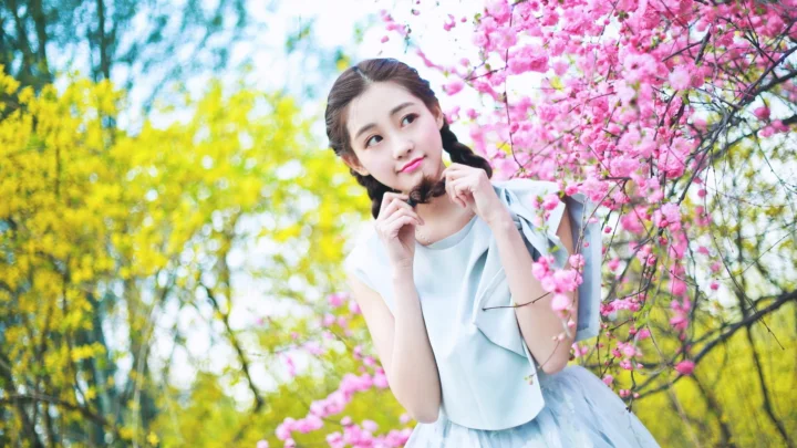 一名年轻女性站在粉色花树旁，周围是明亮的春日树木。