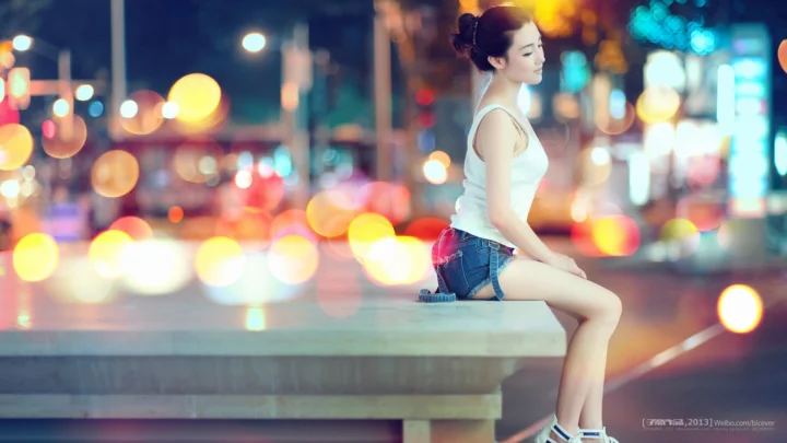 一名成年女子坐在城市夜景前的台面上，背景呈现朦胧多彩的灯光散景。