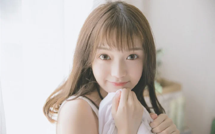 갈색 긴 머리의 여성이 실내의 부드러운 빛 속에서 카메라 가까이 다가와 흰색 천을 두 손으로 가볍게 잡고 있습니다.