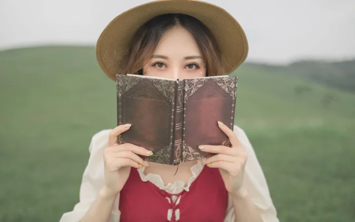챙이 넓은 모자를 쓴 성인 여성이 야외에서 책으로 얼굴 아래쪽을 가리고 있습니다.