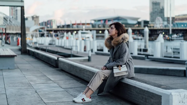 一名女子穿着灰色外套坐在滨水平台边缘，身后是码头和城市建筑。