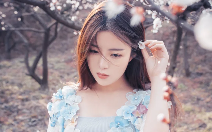 숲속에서 성인 여성이 작은 흰 꽃을 들고 연한 파란색 꽃 장식 의상을 입고 있다.